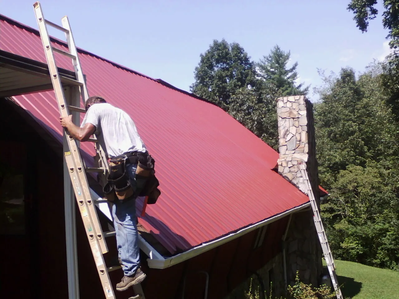 Roofer on a ladder beside a red metal roof and stone chimney — Soffit & Fascia Repair by Roofing Brothers in Chebeague Island